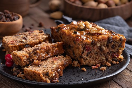 Fruitcake loaf sliced on a plate with raisins and nuts in background on a rustic wooden table